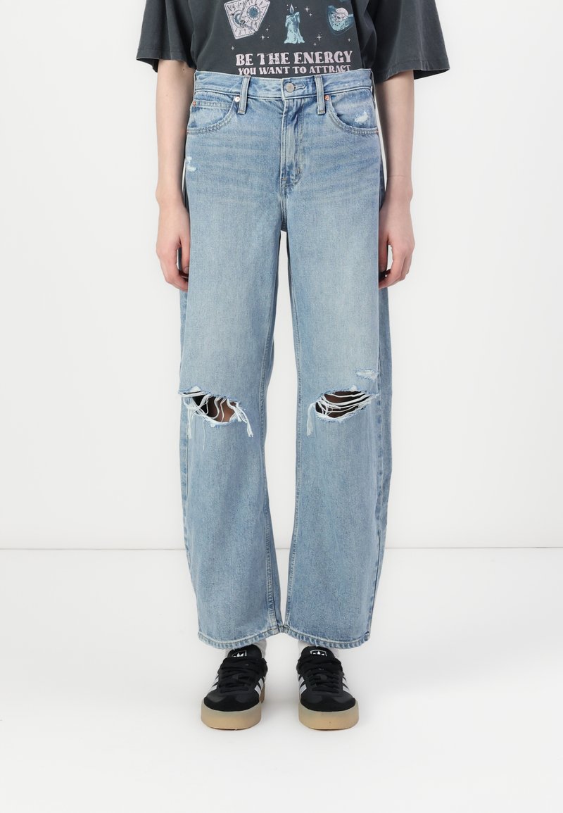 Jean taille haute bleu clair avec jambes larges et déchirures aux genoux. Porté avec des baskets noires ornées de rayures blanches.