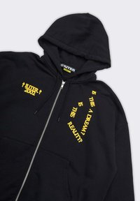 Felpa con zip nera realizzata in cotone, con testo stampato giallo di dimensioni e orientamenti variabili, inclusi il nome del marchio e una domanda.