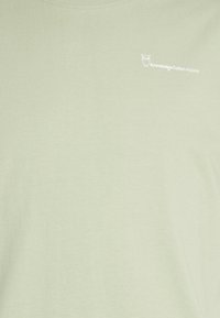 Světle zelená látka s malým bílým logem sovy a textem "Knowledge Cotton Apparel" v horním levém rohu.