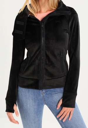 Kobieta ubrana w czarną polarową kurtkę na zamek z otworami na kciuki i dużą klapą na ramieniu, w połączeniu z jasnoniebieskimi dżinsami.