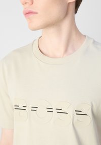 Beidzs krāsas kokvilnas T-krekls ar izteiktu "BOSS" logotipu reljefos, slāņotajos burtiem, akcentēts ar plānām melnām svītrām. Apaļā kakla dizains.