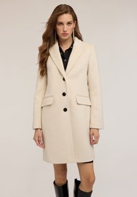 Donna che indossa un cappotto beige al ginocchio con bottoni neri sopra un completo nero, in piedi su uno sfondo neutro chiaro.
