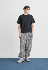 Czarny t-shirt z krótkim rękawem, luźne spodnie w białe i czarne wzory oraz szare trampki, stojące na niebieskiej powierzchni na jednolitym tle.