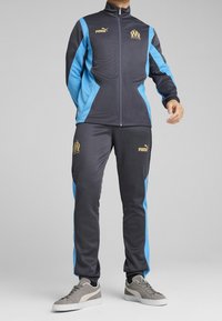 Chándal negro y azul con chaqueta de cremallera y pantalones a juego. Presenta logos y detalles dorados, confeccionado con un material suave y atlético.