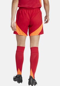 Rode shorts met oranje accenten en een geometrisch ontwerp, gecombineerd met kniekousen in het rood met oranje driehoekige patronen. Zwarte voetbalschoenen zijn zichtbaar.