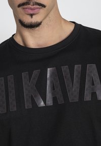 Camiseta de algodón negra con letras grandes y brillantes en negro con un patrón de cuadrícula que dice "KAVA" en el pecho. Cuello redondo estándar.