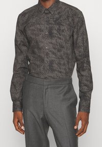 Chemise à manches longues en tissu gris foncé avec un motif tacheté noir subtil. Dotée d'une patte boutonnée à l'avant et d'un col classique. Associée à un pantalon gris.