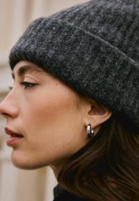 Berretto grigio lavorato a maglia con una texture a coste, abbinato a un orecchino a cerchio nero lucido. Capelli castani, con una finitura liscia e naturale.