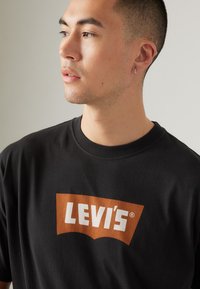 Čierny bavlnený tričko s okrúhlym výstrihom, s veľkým oranžovým a bielym logom Levi's v zakrivenom dizajne na prednej strane. Hladká textúra.