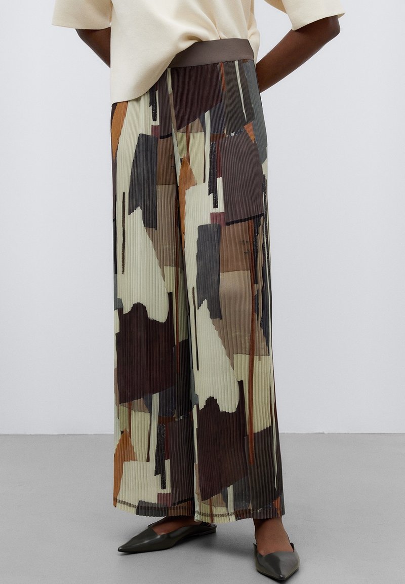 Femme portant un pantalon plissé à jambes larges avec des motifs abstraits marron, beige et noir, associé à des ballerines pointues foncées.