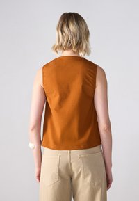 Ærmeløs top i rustorange stof, med en rund halsudskæring og en glat tekstur. Sammen med lyse beige højtaljede bukser og baglommer.
