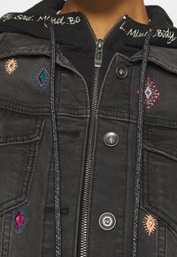 Svart denimjacka med broderade diamantmönster i rosa, turkos och gult. Har en dragkedja framtill och dubbla tryckknappar.