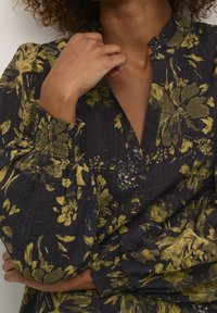 Blusa floral con fondo oscuro, que presenta un patrón de flores amarillas y verdes, un escote en V, mangas fruncidas y un tejido texturizado.