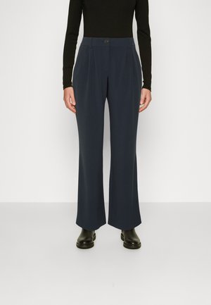 Femme portant un pantalon large bleu marine foncé taille haute, un haut noir à manches longues et des bottines noires, debout devant un fond blanc.