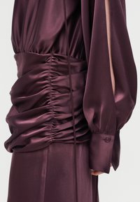 Μακρύ φόρεμα από βαθύ μοβ σατέν με σφηκοφωλιά στη μέση, μακριά φουσκωτά μανίκια με λεπτομέρειες σχισμών και μανσέτες με κουμπιά.