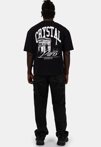 Czarna bawełniana koszulka z białym nadrukiem graficznym na plecach, przedstawiająca "CRYSTAL Paris" oraz ilustrację architektoniczną. Założona z czarnymi spodniami cargo.