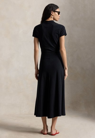 Žena v čiernych midi šatách s krátkym rukávom, čiernych slnečných okuliaroch, zlatých kruhových náušniciach a červených plochých sandáloch, stojaca pred jednoduchým pozadím.