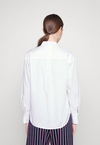 Camicia bianca a maniche lunghe con yoke posteriore e sottili spacchi sui lati. Tessuto liscio, polsini con bottone e orlo arcuato. Abbinata a pantaloni a righe.