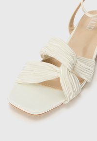 Sandal en satin blanc avec un bout carré, présentant des brides plissées croisées et une doublure intérieure beige. Son design est minimaliste et sa texture lisse.