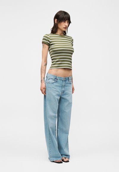 Γυναίκα με τατουάζ που φοράει πράσινη και λευκή ριγέ κοντή μπλούζα, ανοιχτόχρωμο τζιν παντελόνι με φαρδιά μπατζάκια και μαύρα σανδάλια ανοιχτά στα δάχτυλα, στέκεται μπροστά σε λευκό φόντο.