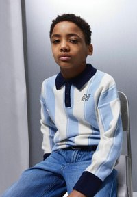 Poiss istub toolil, seljas valge ja helesinise triibuline kampsun tumedate mansettide ja kraega, koos siniste teksadega.