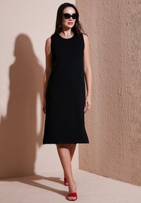 Black sleeveless dress with a relaxed fit, smooth fabric, knee-length hem, paired with red heeled sandals and dark sunglasses.