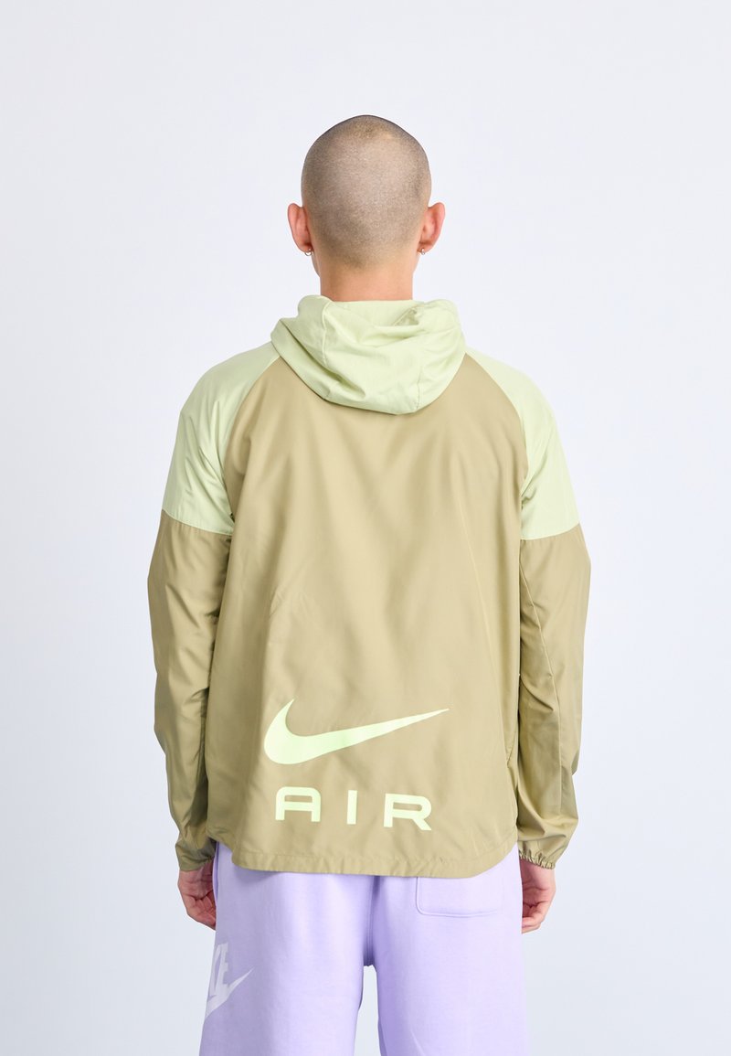Beige jas met een capuchon, lichtgroene accenten en een groot wit Nike-logo op de achterkant. Gladde textuur, lichtgewicht materiaal.