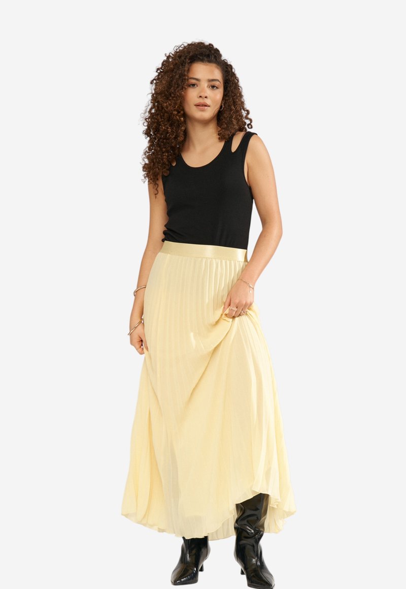 Femme aux cheveux bouclés portant un haut noir sans manches, une jupe plissée jaune pâle et des bottes noires à talons, tenant la jupe d'une main.