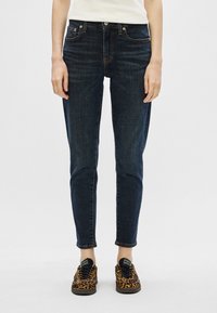 Granatowe dżinsy o slim fit z pięcioma kieszeniami, z kontrastowym złotym przeszyciem, noszone z trampkami w leopardzi wzór.