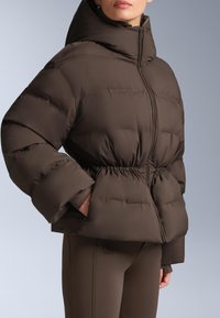 Smeđa puhasta jakna s visokim ovratnikom, elastičnim strukom i prevelikim rukavima. Izrađena od glatkog, teksturiranog materijala.