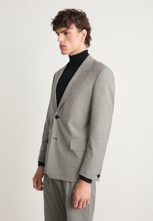 Kohandatud hall ruuduline ülikond ühe nupu sulgemisega, klappidele ja küljetaskutele. Kandarüüs must turtleneck. Tekstuuriga kangas.