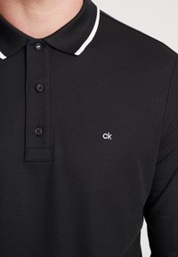 Svart polo skjorta med strukturerad textil, med en treknapps knäppning, vit krage med rand och en liten vit logotyp på bröstet.