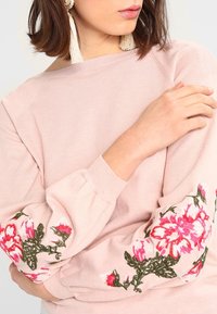 Hellrosa Pullover mit gerippter Struktur und floraler Stickerei in Pink und Grün auf den Ärmeln. Das Design zeichnet sich durch einen weiten Ausschnitt und Puffärmel aus.