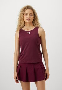 Mujer con pelo rubio rizado, vistiendo una camiseta deportiva sin mangas de color vino y una falda plisada a juego, de pie frente a un fondo liso.