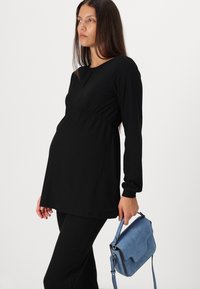 Schwarzes, geripptes Langarmoberteil mit figurbetonter Silhouette und Rundhalsausschnitt, kombiniert mit schwarzen Hosen. Eine hellblaue Wildleder-Umhängetasche wird gehalten.