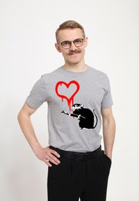 Šedé tričko s krátkým rukávem má grafiku černé krysy s červeným srdcem, ze kterého kapají kapky. Model nosí černé kalhoty, ruce v bok.