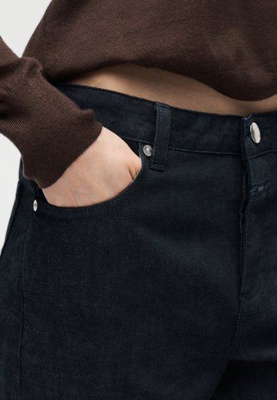 Κοντινό πλάνο ενός ατόμου που φοράει σκούρο μπλε τζιν, με το ένα χέρι στην μπροστινή τσέπη και ένα καφέ πουλόβερ με μακριά μανίκια.