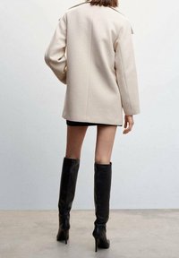 Blazer oversize beige con un design strutturato, in tessuto misto lana e dettagli sulle spalle inclinati. Abbinato a stivali neri alti fino al ginocchio con tacco a stiletto.