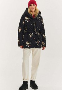 Čierna prešívaná bunda s kvetinovými vzormi, kapucňou a opaskom; v kombinácii s krémovými nohavicami a čiernymi čižmami. Teplá, textúrovaná látka.