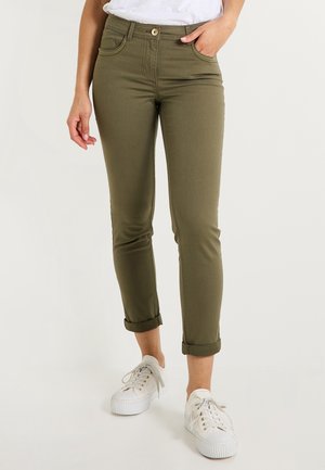 Femme portant un pantalon vert olive retroussé, des baskets blanches et un haut blanc avec une main dans la poche.