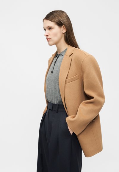 Nő teveszínű kabátban, szürke garbóingben és magas derekú fekete nadrágban, zsebében tartva a kezét, balra néz egy egyszínű háttér előtt.