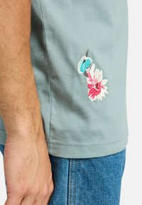 Lys blå bomulds t-shirt med blomsterbroderi i pink, hvid og turkis på venstre side. Klassisk rund halsudskæring og korte ærmer.
