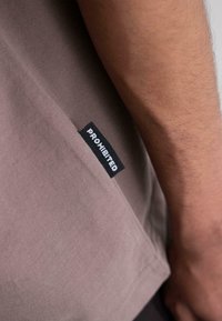 Brown short-sleeve T-shirt with a textured fabric and a black tag reading "PROHIBITED" on the lower side near the hem.