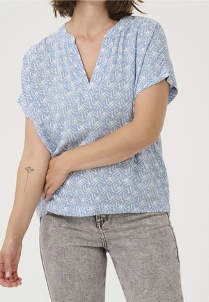 Vrouw die een lichtblauw gebloemd V-halsblouse en lichtgrijze jeans draagt, met een kleine bloemen-tatoeage op haar linker onderarm.