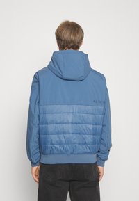 Modrá péřová bunda s kapucí, která má hladkou horní část a žebrovaný pas, a na zádech ukazuje horizontální prošívaný vzor.