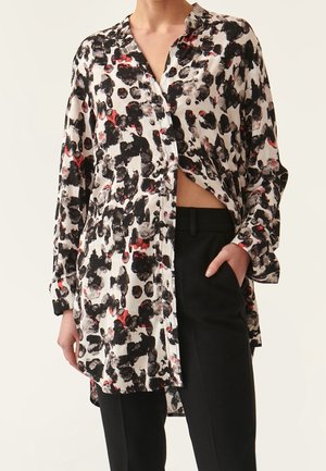 Femme portant un chemisier blanc à manches longues avec des taches abstraites noires et rouges, partiellement déboutonné, et un pantalon noir avec une main dans la poche.