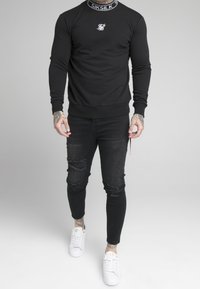 Man som bär en svart Siksilk-tröja, revade svarta skinny jeans, vita Siksilk-sneakers och synliga tatueringar på händer och anklar.