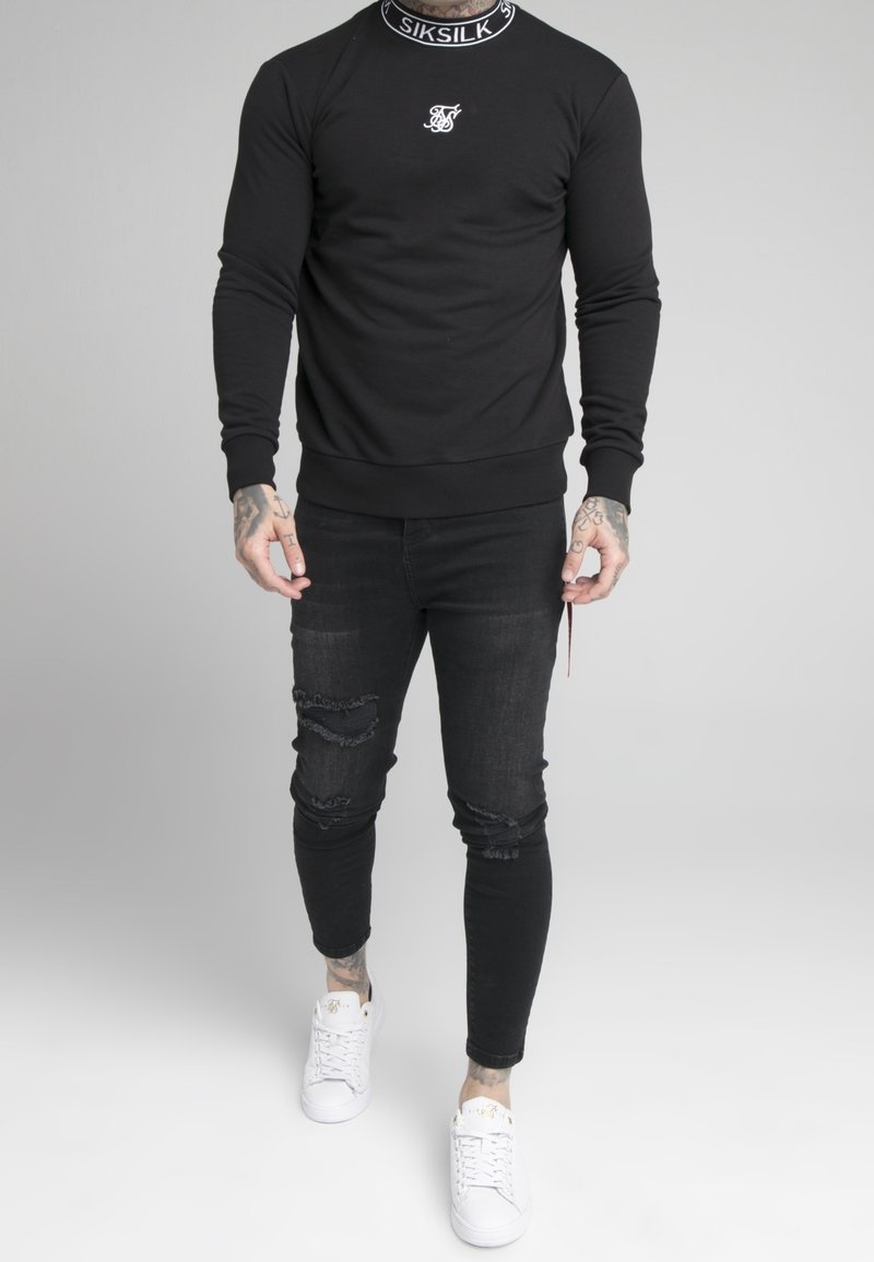 Man som bär en svart Siksilk-tröja, revade svarta skinny jeans, vita Siksilk-sneakers och synliga tatueringar på händer och anklar.