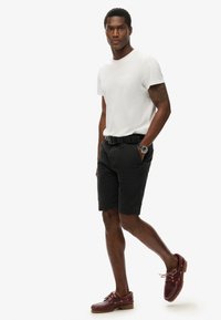 T-shirt bianca a girocollo, pantaloni corti neri, cintura nera e scarpe da barca bordeaux. Orologio notevole al polso, pantaloni corti con tasche, vestibilità casual.