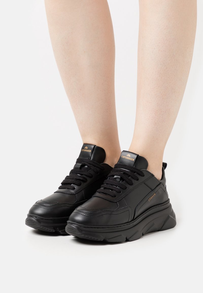 Baskets en cuir noir à lacets avec semelles épaisses portées sur des jambes nues, affichant la marque « Copenhagen » en lettres dorées sur la languette et sur le côté.